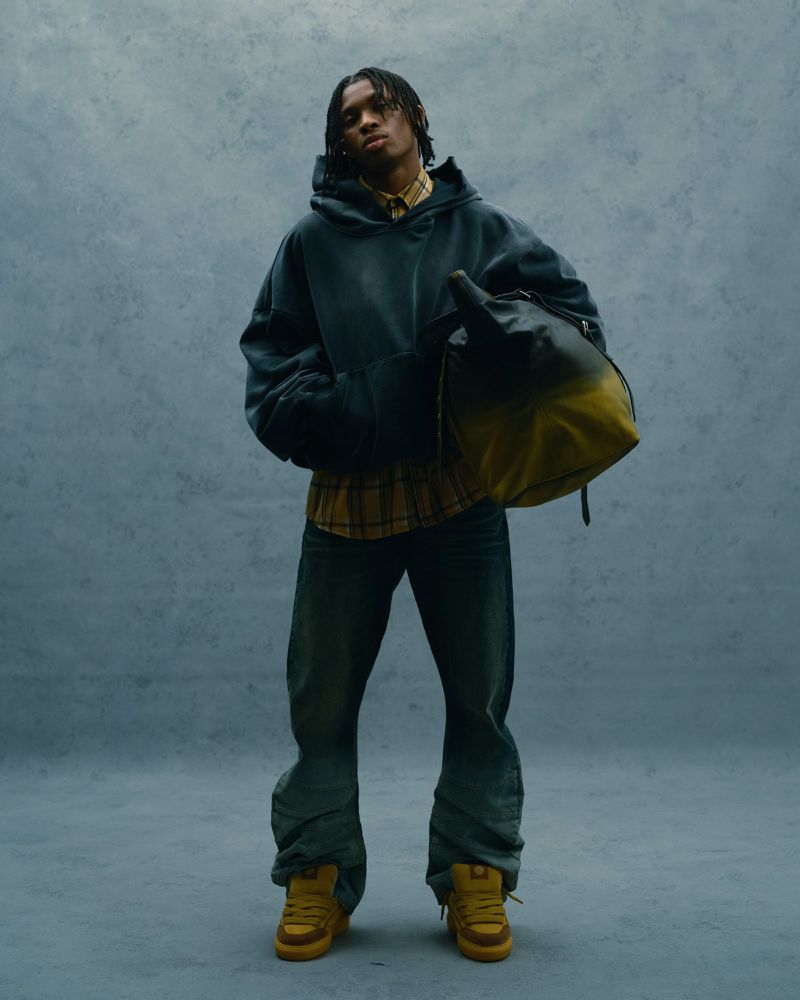 Young adult wearing a hoodie and jeans, holding a bag, in a studio shot against a gray backdrop with soft, neutral lighting.