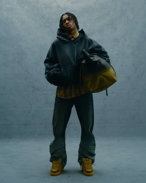 Young adult wearing a hoodie and jeans, holding a bag, in a studio shot against a gray backdrop with soft, neutral lighting.