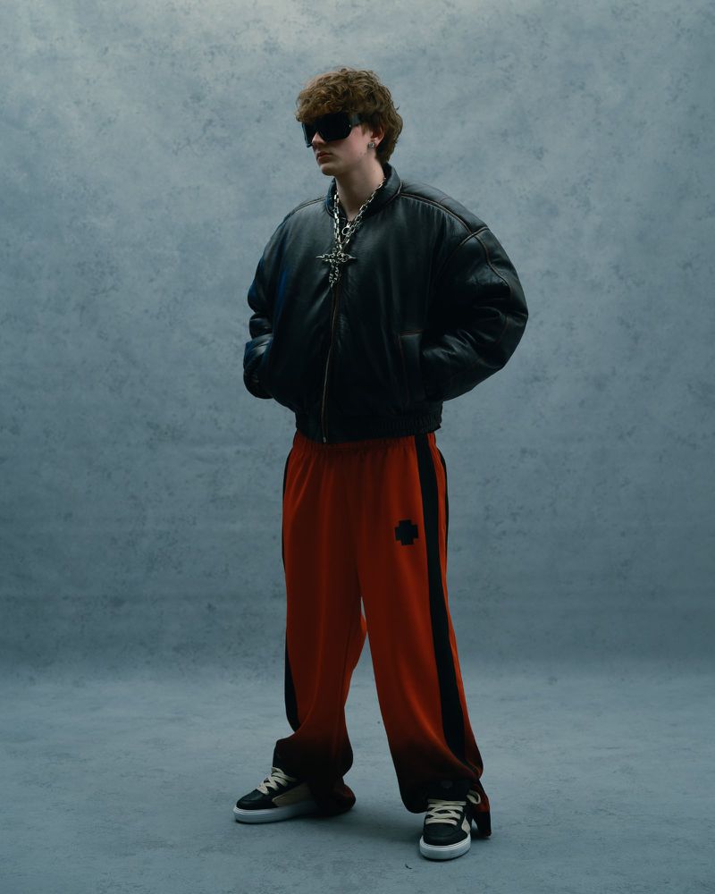Adult male in a black bomber jacket and orange sweatpants stands in a photo studio with gray background in a full shot.