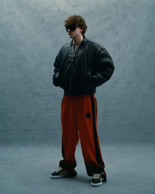 Adult male in a black bomber jacket and orange sweatpants stands in a photo studio with gray background in a full shot.