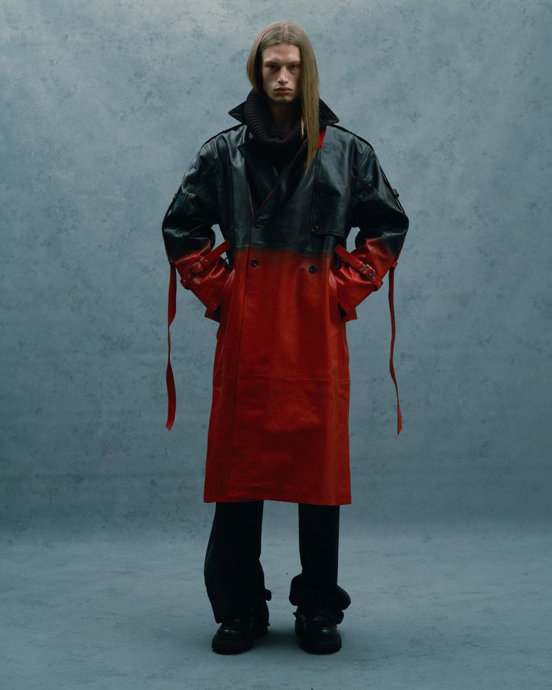 A tall adult male model with long hair poses in a red-and-black leather coat, black pants, and shoes, indoors against a textured grey backdrop.