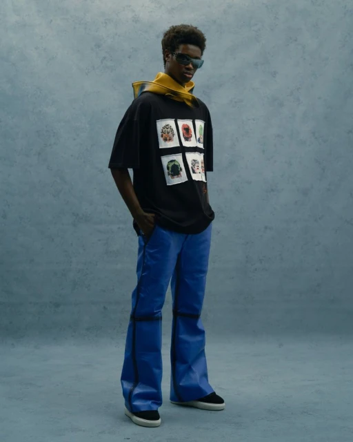 Full-length shot of a Black male model with sunglasses wearing a graphic t-shirt, blue leather pants, and black sneakers in a studio setting.