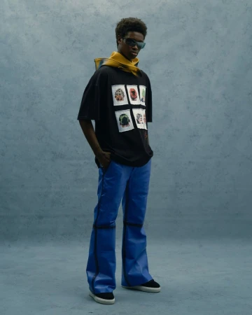 Full-length shot of a Black male model with sunglasses wearing a graphic t-shirt, blue leather pants, and black sneakers in a studio setting.