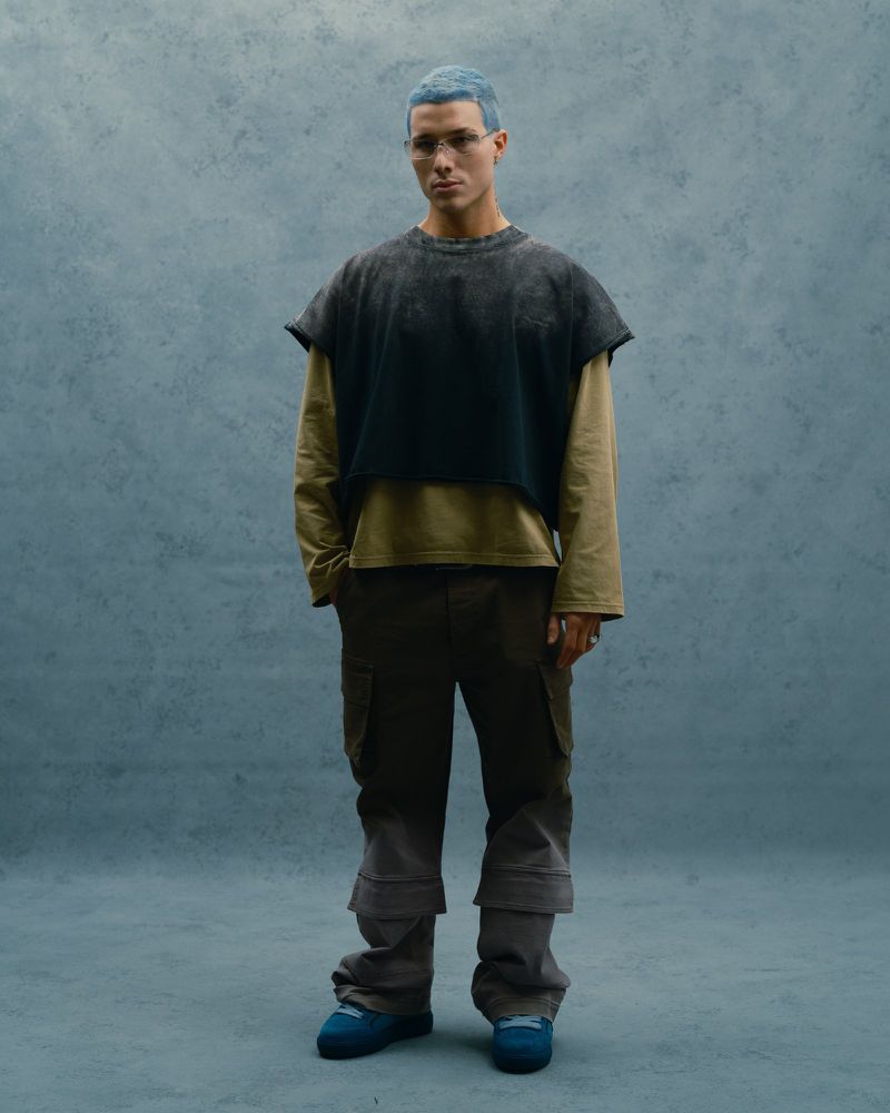 Full-shot of a young adult with light blue hair wearing a dark top over a tan shirt, brown pants, and blue shoes against a blue backdrop.
