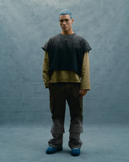Full-shot of a young adult with light blue hair wearing a dark top over a tan shirt, brown pants, and blue shoes against a blue backdrop.