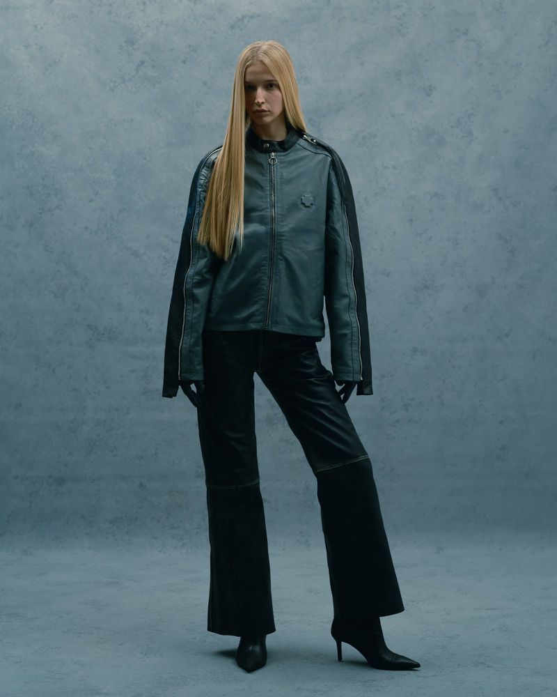 Full-length studio shot of a woman with long blond hair in a grey jacket, black wide-leg pants, gloves, and high-heeled shoes against a grey background.