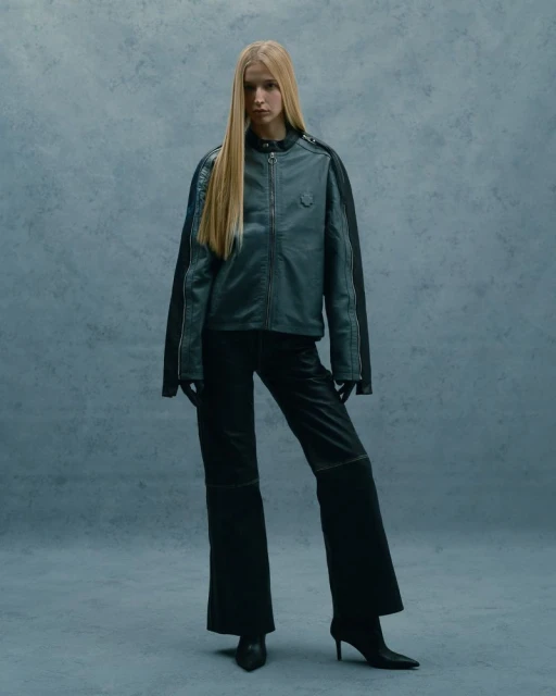 Full-length studio shot of a woman with long blond hair in a grey jacket, black wide-leg pants, gloves, and high-heeled shoes against a grey background.
