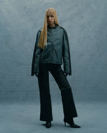 Full-length studio shot of a woman with long blond hair in a grey jacket, black wide-leg pants, gloves, and high-heeled shoes against a grey background.