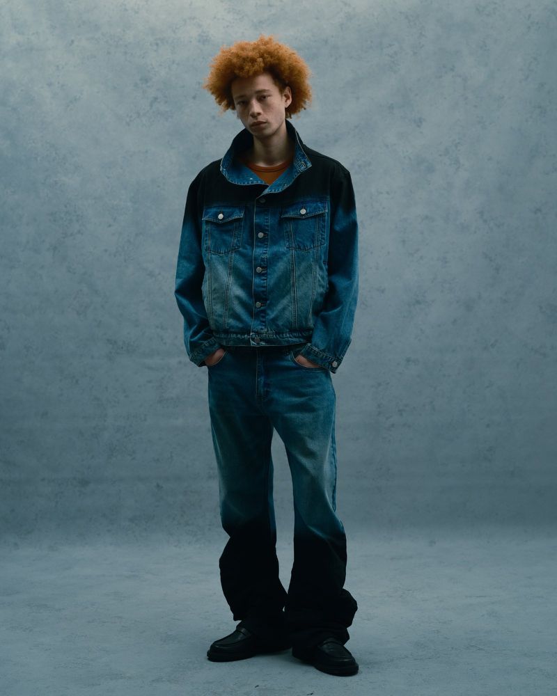 Young adult with orange curly hair wears a faded blue denim jacket and jeans against a textured blue photo studio backdrop.