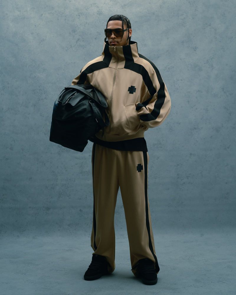 Man in a beige tracksuit and sunglasses stands holding a black backpack against a muted blue background in a full-length fashion shot.