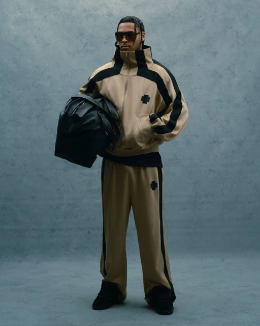 Man in a beige tracksuit and sunglasses stands holding a black backpack against a muted blue background in a full-length fashion shot.