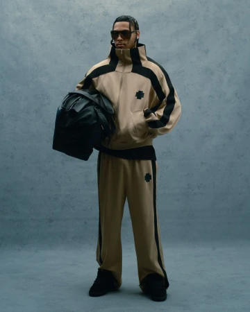 Man in a beige tracksuit and sunglasses stands holding a black backpack against a muted blue background in a full-length fashion shot.