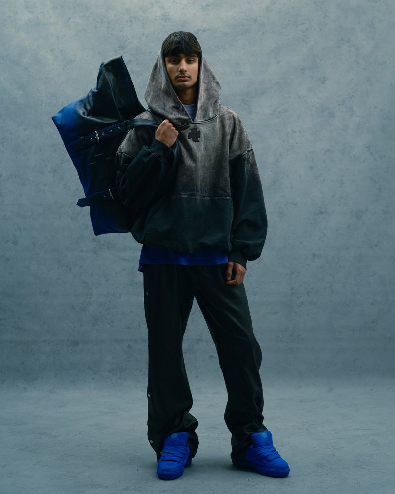 Man in grey ombre hoodie, dark trousers, and bright blue shoes standing with duffel bag against mottled grey studio backdrop.