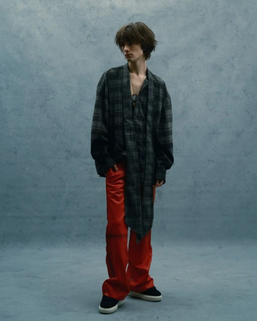 Full-length shot of a young adult in a grey plaid shirt, red satin pants, and black shoes, standing against a textured blue background.