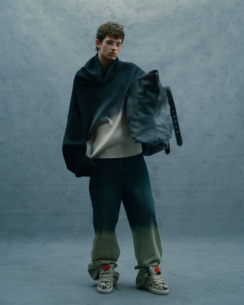 Model wearing a gradient sweater and dark trousers holding a large bag against a textured grey background.