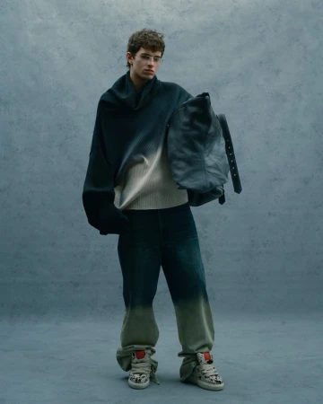 Model wearing a gradient sweater and dark trousers holding a large bag against a textured grey background.