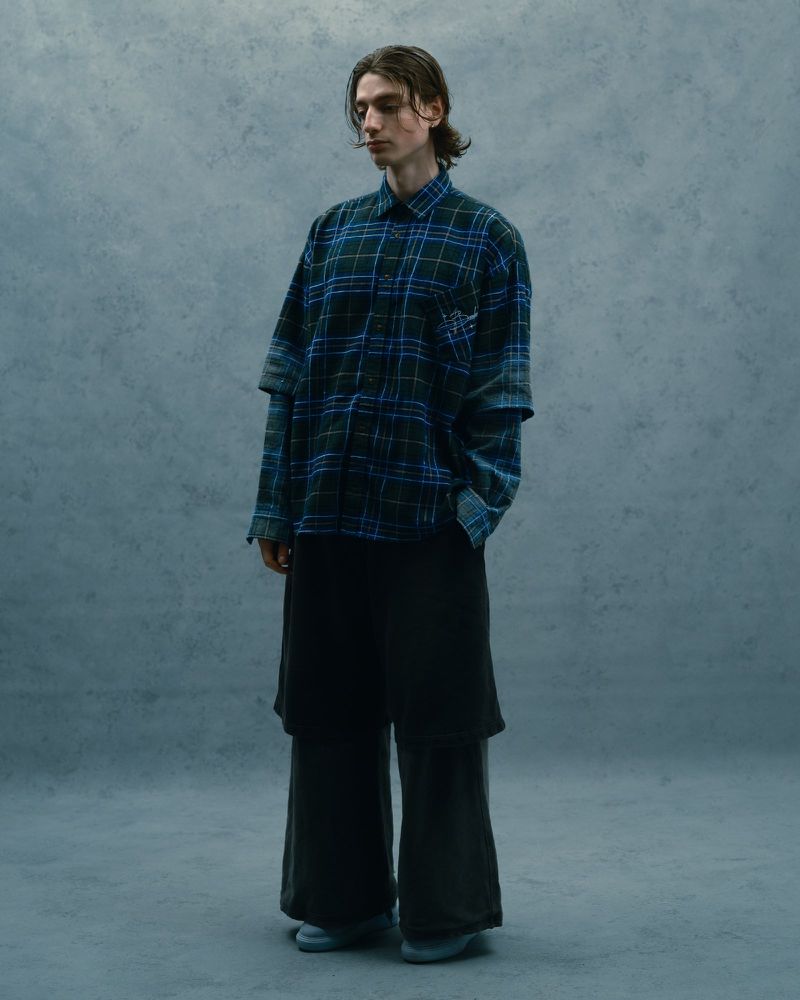 Full shot of a young adult man with medium-length brown hair wearing a plaid shirt and dark pants in a studio setting with grey backdrop.