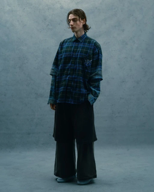 Full shot of a young adult man with medium-length brown hair wearing a plaid shirt and dark pants in a studio setting with grey backdrop.
