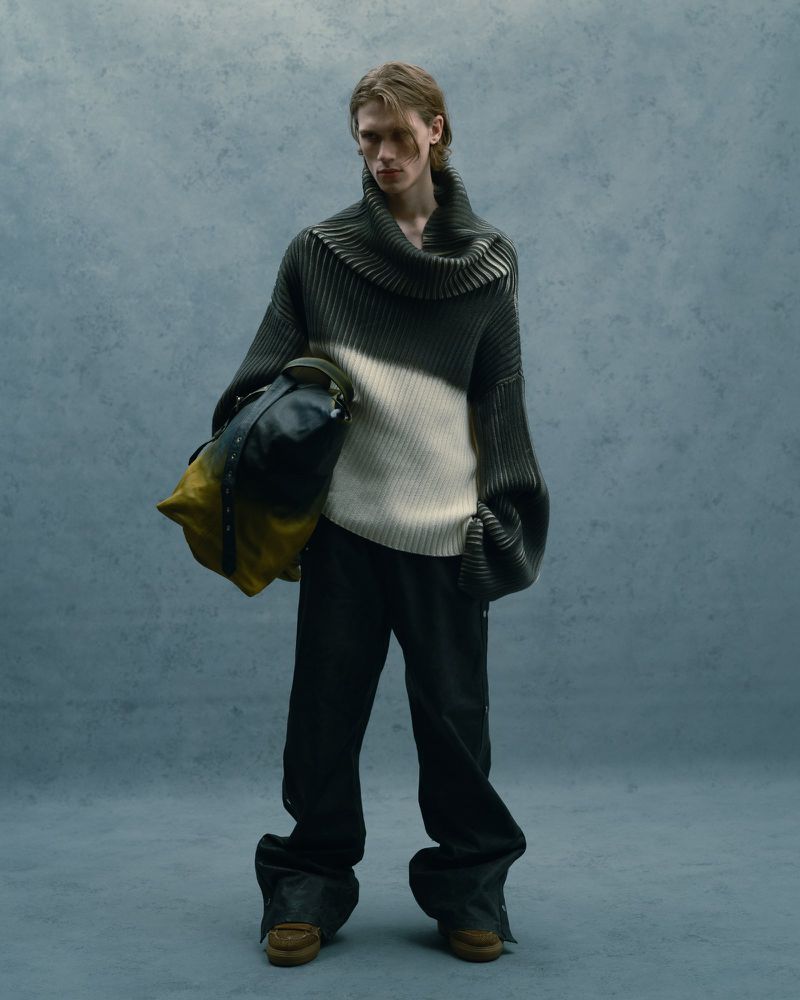 Male fashion model poses in an indoor setting with a grey and white sweater, dark pants, and carries a duffel bag.