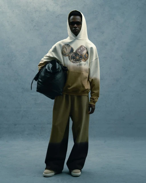 Man in hoodie and sweatpants holds black bag in studio shot with neutral backdrop, showcasing casual fashion apparel.
