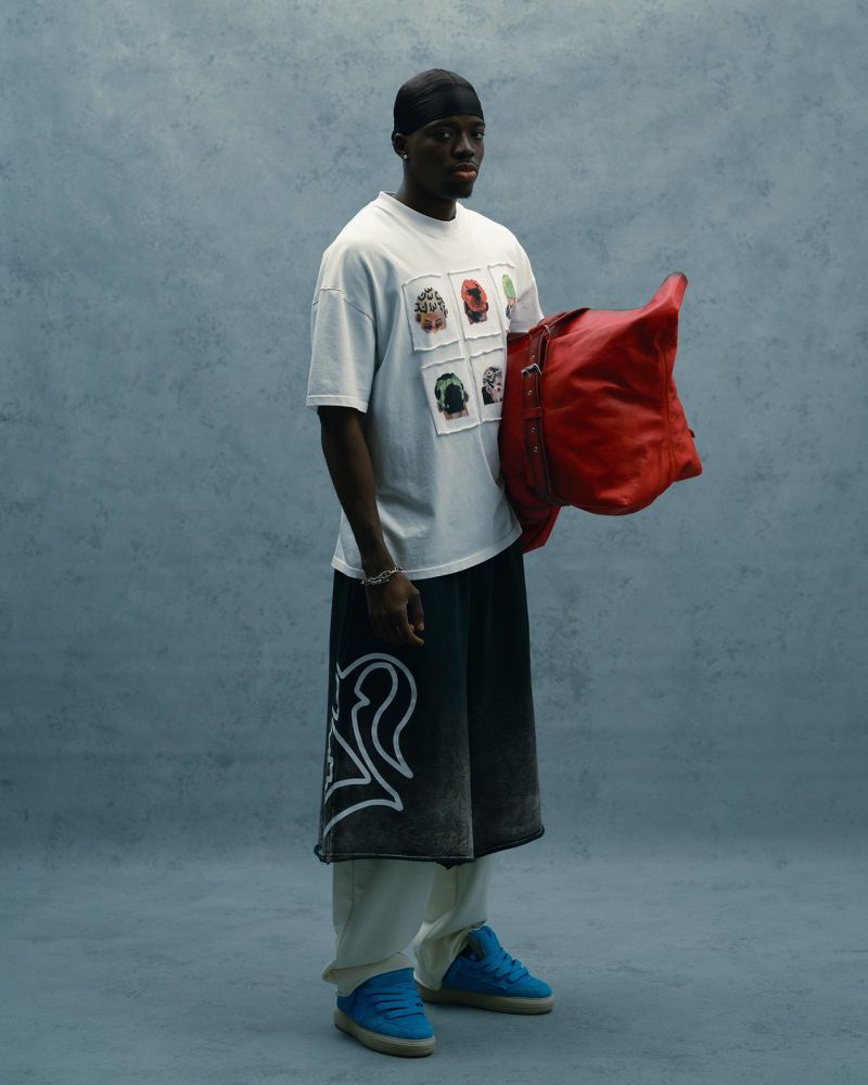 A person in an oversized graphic tee, shorts, and sneakers stands against a mottled blue backdrop, holding a red duffel bag.