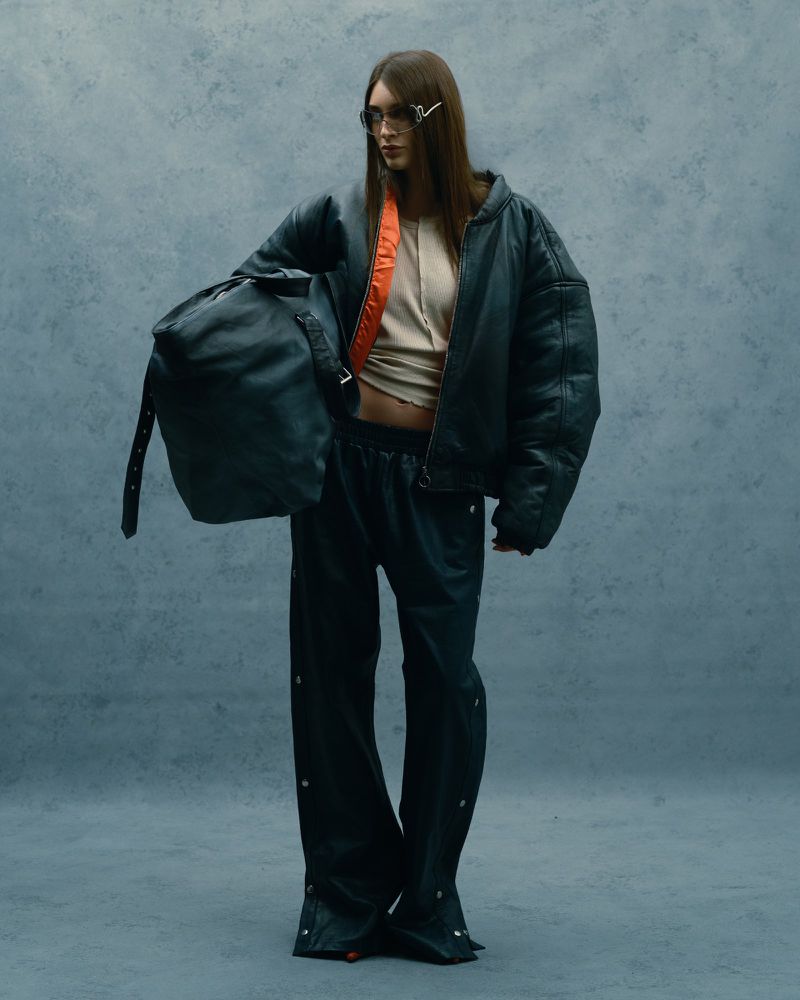 A woman in a dark oversized bomber jacket, sunglasses, and baggy pants, holding a duffle bag, stands in a neutral-toned photo studio.