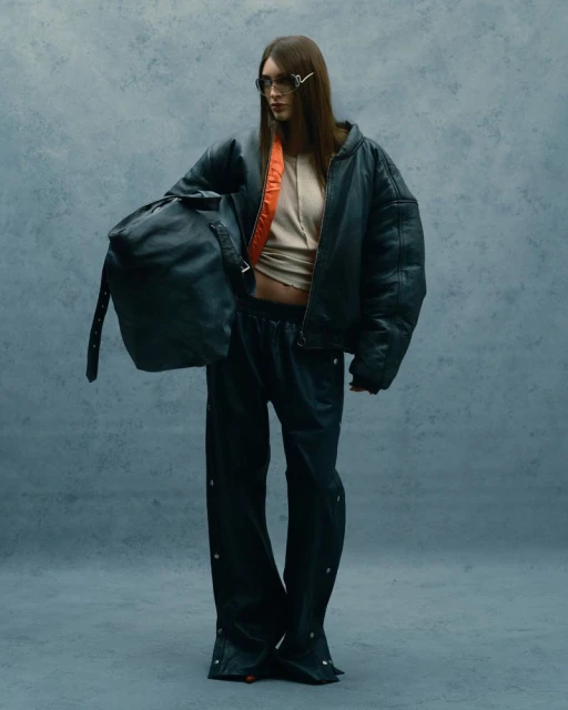 A woman in a dark oversized bomber jacket, sunglasses, and baggy pants, holding a duffle bag, stands in a neutral-toned photo studio.