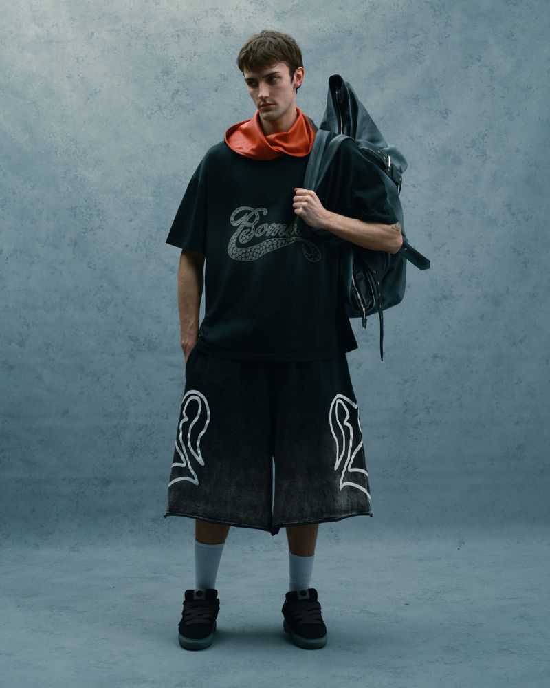 Full shot of a young adult man in streetwear-style clothing, featuring a t-shirt, shorts with graphic designs, sneakers, and a backpack.