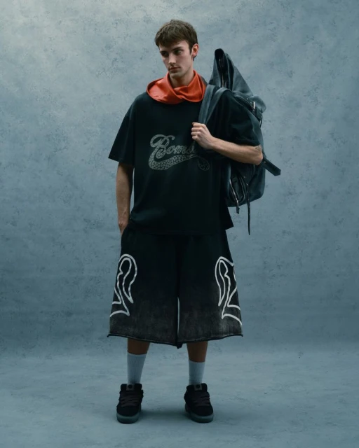 Full shot of a young adult man in streetwear-style clothing, featuring a t-shirt, shorts with graphic designs, sneakers, and a backpack.