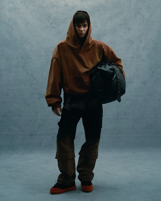 Model in a brown hooded sweatshirt holding a black leather duffel bag against a textured grey studio backdrop