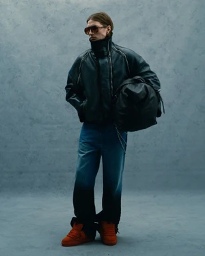Model in dark leather jacket, sunglasses, and orange sneakers carrying a large bag against a mottled grey wall.