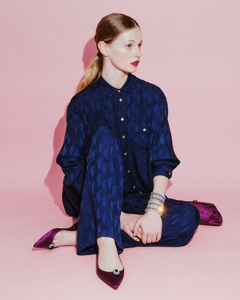 A young fair-skinned woman in a patterned navy outfit sitting cross-legged on a pink background with pointed purple shoes and jewelry.