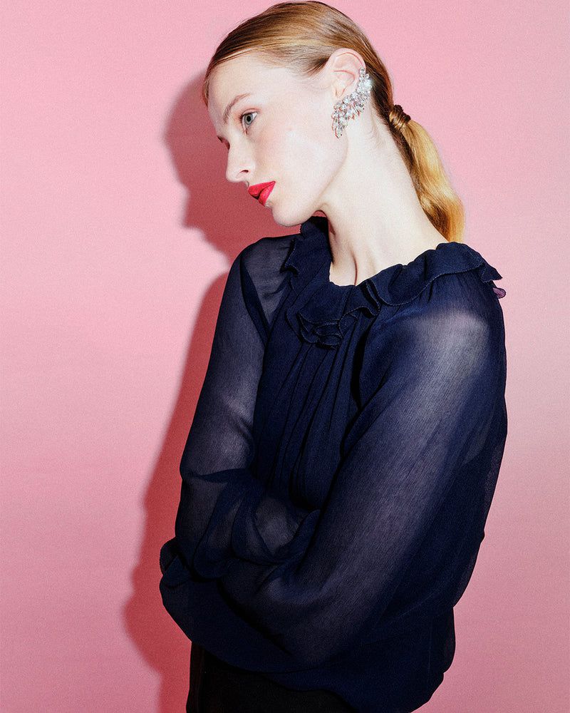 Elegant woman with red lipstick and a sheer navy blouse poses against a pink background, highlighting her sophisticated style and fashion.