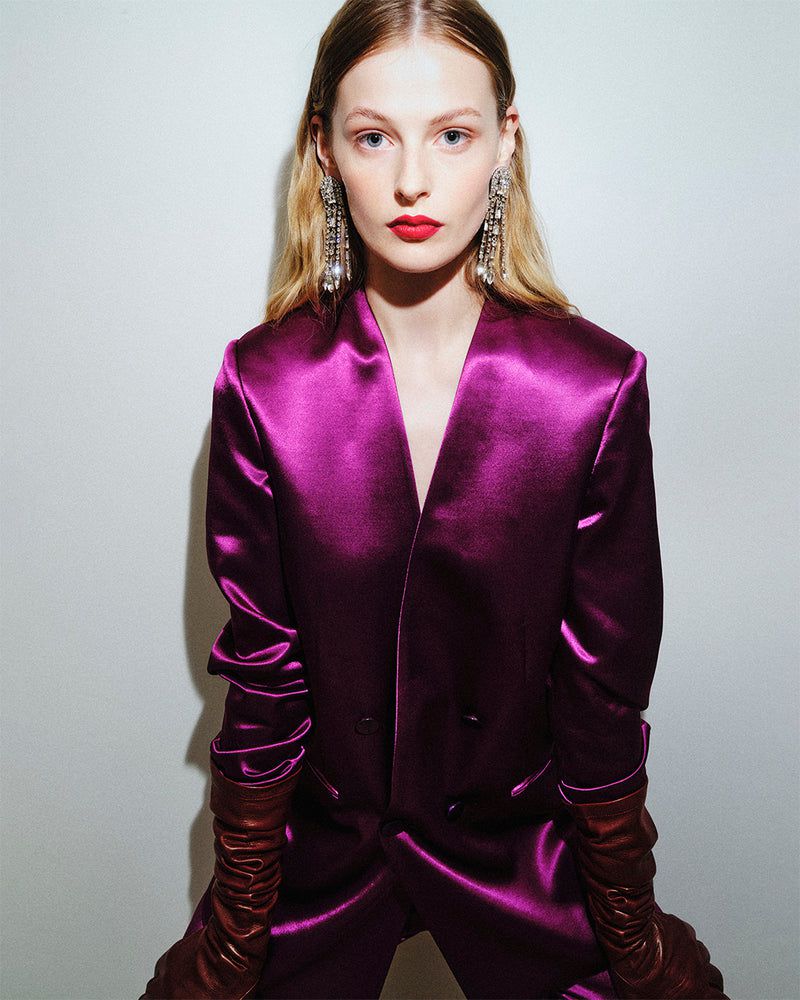 Woman with blonde hair in a vibrant magenta satin suit with long gloves posing against a plain white background, wearing chandelier earrings.