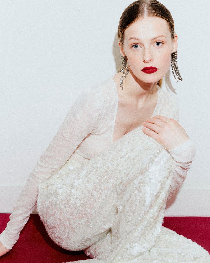 Portrait of a fair-skinned adult woman with red lipstick and statement earrings, wearing a white long-sleeved top and white sequined pants.