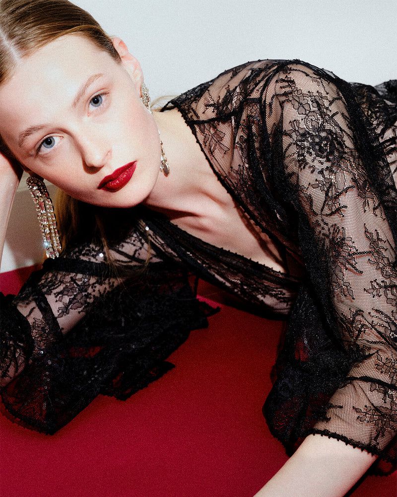 Portrait of a fair-skinned young woman with dark red lipstick wearing a black lace top and dangling earrings on a red surface.