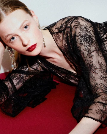 Portrait of a fair-skinned young woman with dark red lipstick wearing a black lace top and dangling earrings on a red surface.
