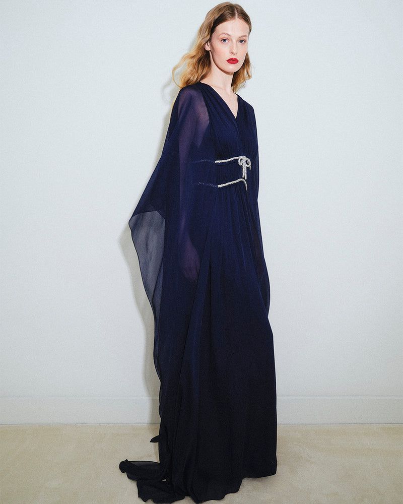 Elegant woman in a flowing navy gown with a jeweled belt, posing against a plain backdrop in a fashion editorial style.