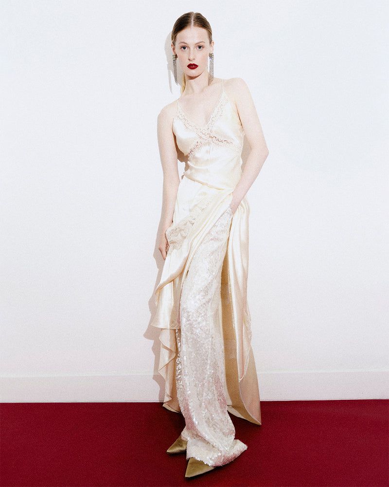 A woman in a gown poses in front of a white wall with a red base. She wears dangly earrings, and her hair is pulled back.