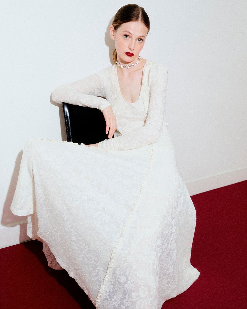 Elegant woman in an off-white gown poses seated on a black chair against a white backdrop with red carpet.