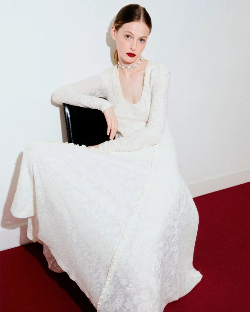 Elegant woman in an off-white gown poses seated on a black chair against a white backdrop with red carpet.