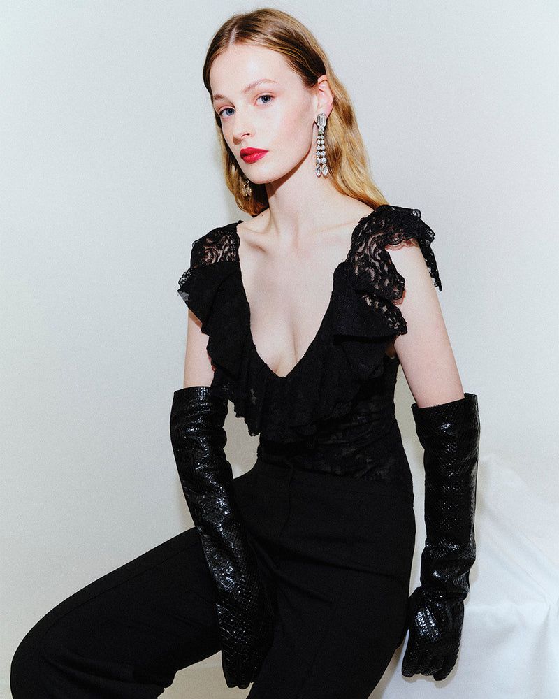 Woman with blonde hair in black lace top and pants, wearing long black gloves and chandelier earrings against a white backdrop.