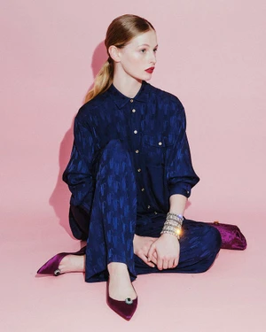 Model seated on a pink floor wearing a dark blue patterned silk shirt and matching trousers with purple pointed heels