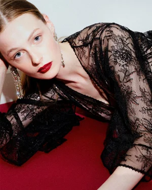 Model lying down wearing a sheer black lace dress with crystal earrings and dark red lipstick on a red surface