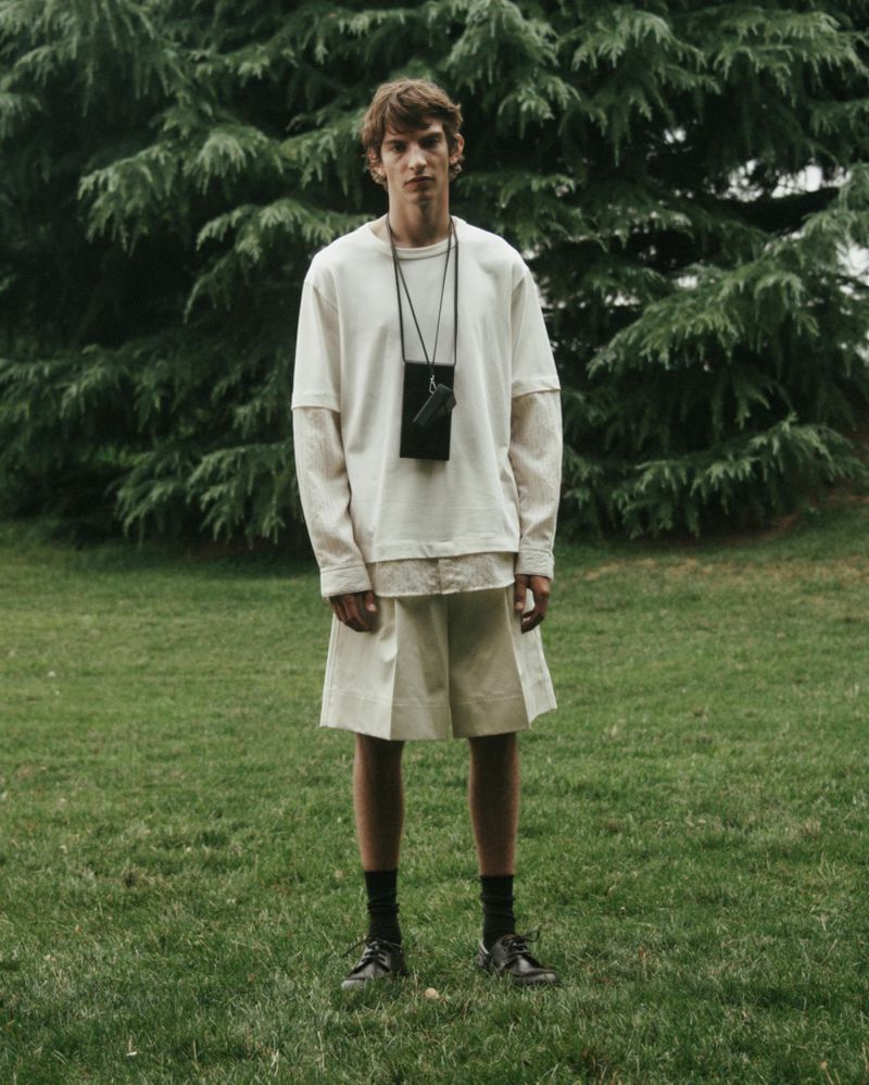 Full-length shot of a male fashion model in an off-white tee shirt and shorts outdoors, posed in a field with trees in the background.