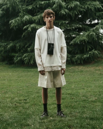 Full-length shot of a male fashion model in an off-white tee shirt and shorts outdoors, posed in a field with trees in the background.