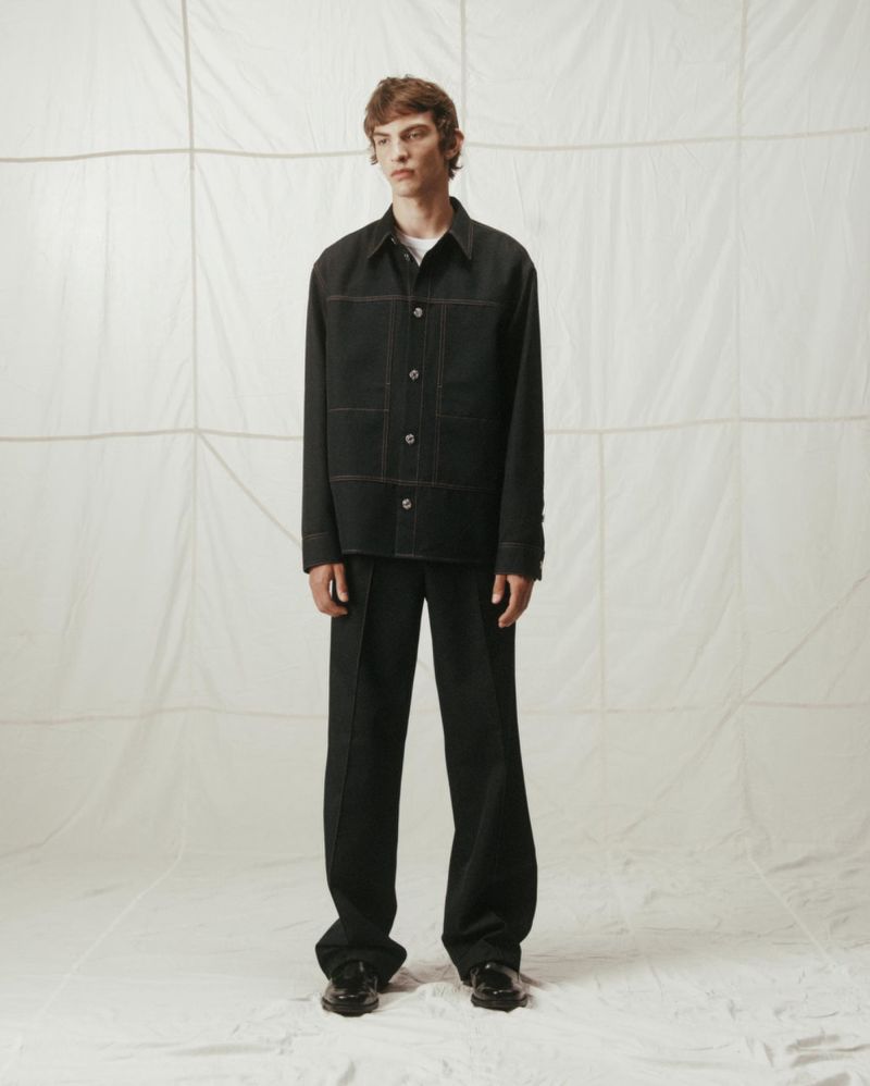 Full-length portrait of a young adult man in a black outfit and dress shoes standing in front of a white background, studio lighting.