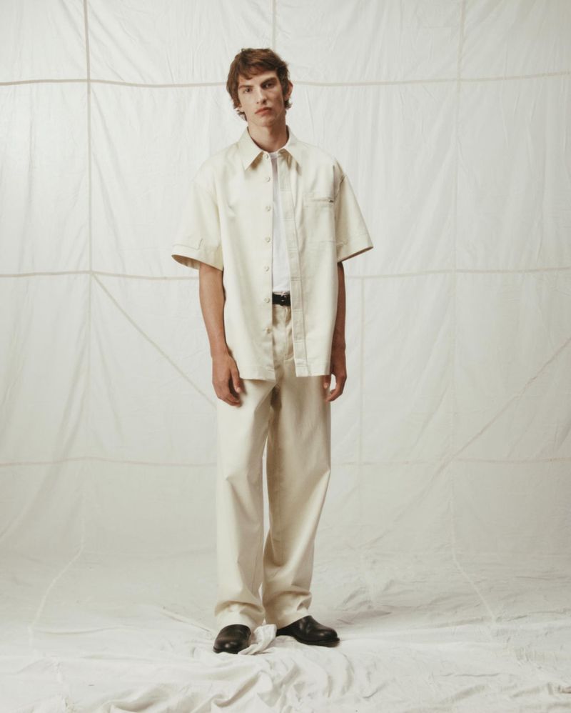 An adult male stands in a photo studio wearing a light beige short-sleeve button-up shirt, cream-colored slacks, and black dress shoes, fashion shoot.