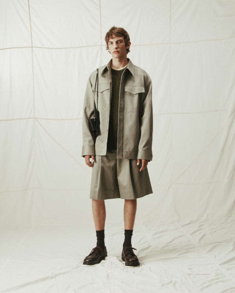 Fashion portrait of a young adult man in a studio wearing a khaki jacket and skirt with brown shoes and socks against a neutral background.