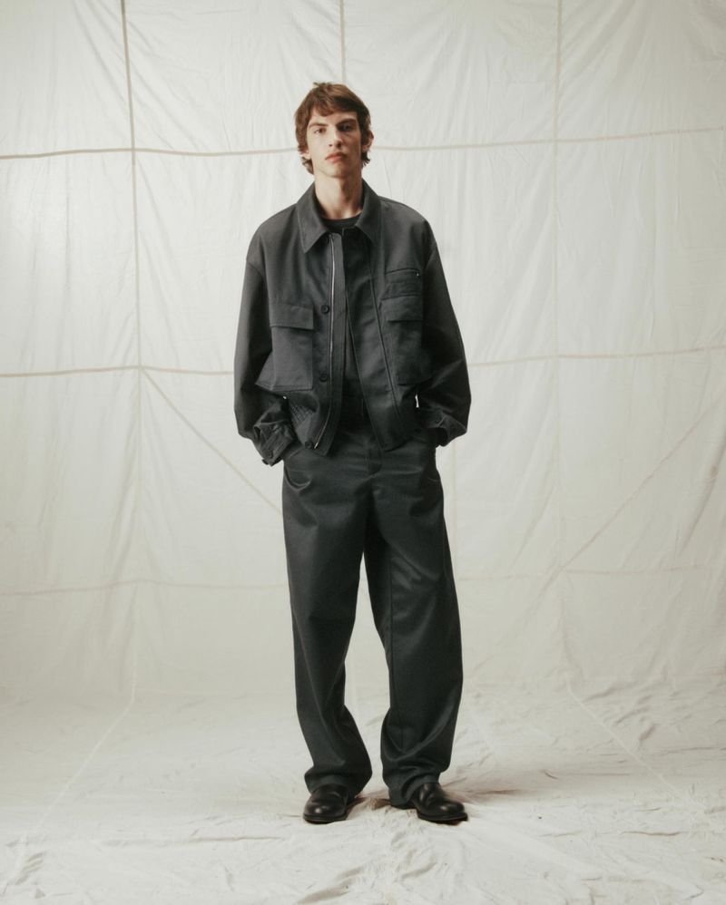 Adult male posing in a dark gray shirt and pants in a photo studio against a white backdrop; fashion editorial, studio lighting.